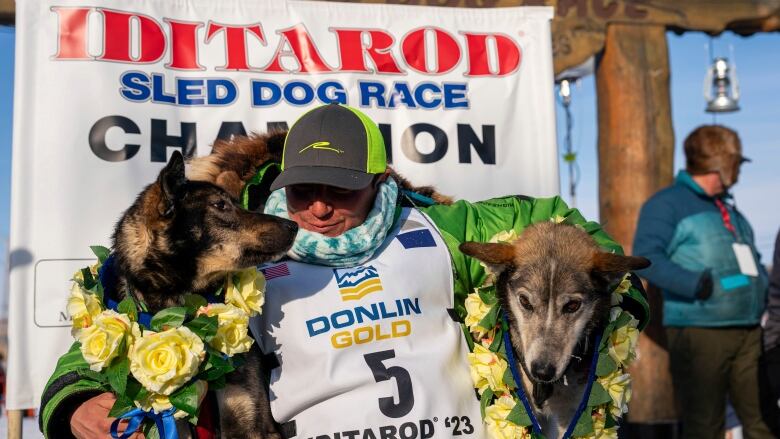 A man poses with his arms around 2 dogs, in front of a banner reading, 'Iditarod Sled Dog Race Champion.'