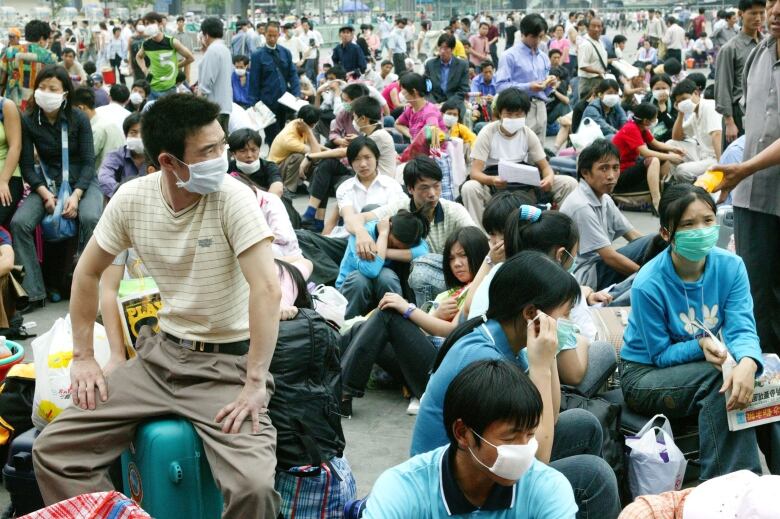 A large crowd of people, some wearing surgical masks, stand or sit in a large open area. A man in the left foreground sits on his luggage.