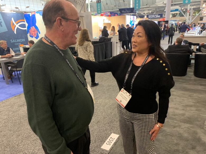 A man in a green shirt speaks with a woman on the sales floor of a conference in Boston. The woman is reaching across and touching the man on his shoulder as they chat. 