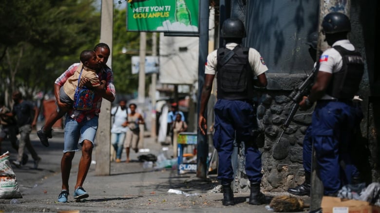 A parent, carrying his child after picking him up from school, runs past police as they carry out an operation against gangs in the Bel-Air area of Port-au-Prince, Haiti, Friday, March 3, 2023. (AP Photo/Odelyn Joseph)