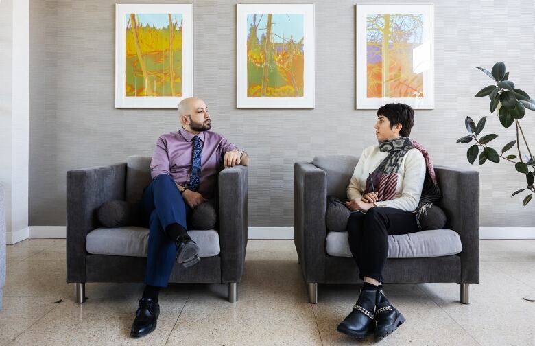 A man and woman sit on two chairs. They look at each other with serious expressions on their faces.