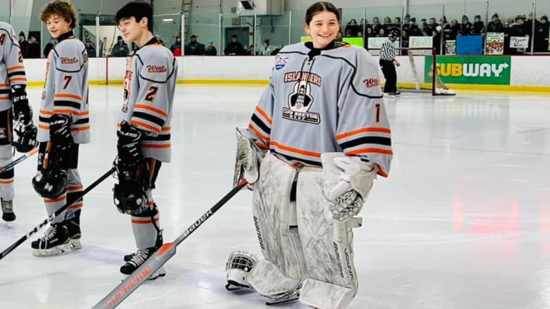 A hockey player wearing goalie pads and a white and orange jersey stands on the ice.