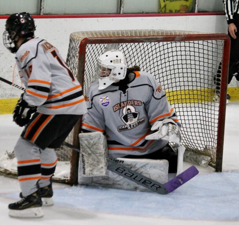 A goalie in a white helmet and white hockey jersey watches the play from the net.