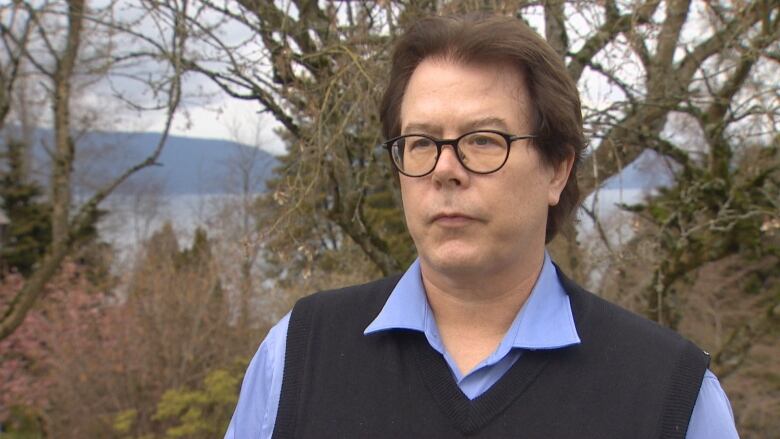 A man wearing a blue shirt and a vest stands in front of a tree on the University of British Columbia campus.