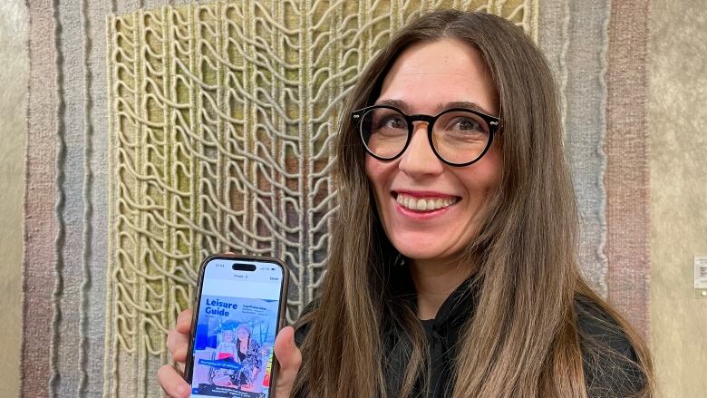 A woman with long brown hair and wearing glasses shows a photo on her mobile phone of magazine cover. 