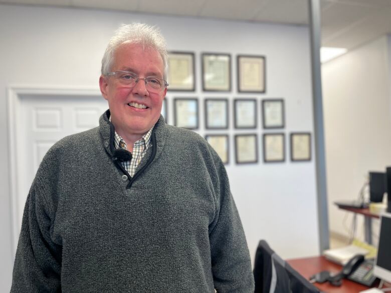 A man in a grey sweater smiles at the camera in front of a wall of certificates.