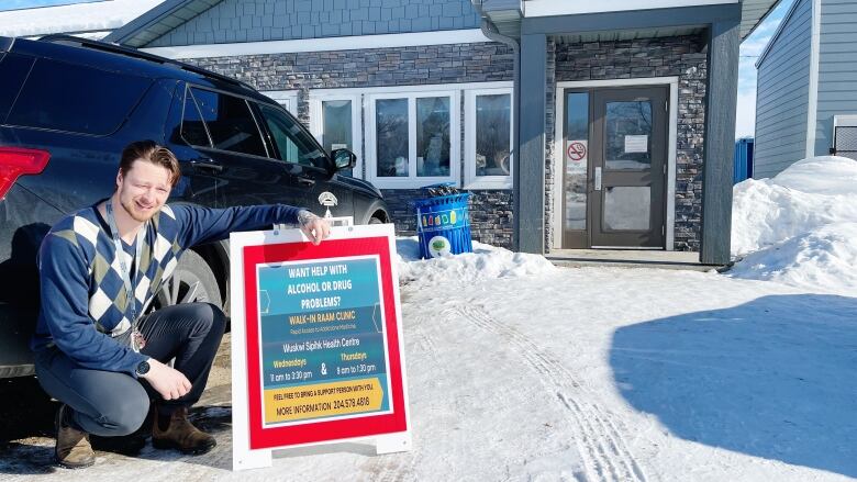 Man with brown hair squats in front of a car. He is leaning on a sign. and wears a blue sweater. 