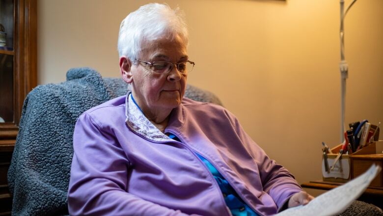 An elderly woman is pictured sitting on a chair and looking down at pieces of paper in her hands.