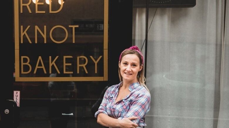 A woman, wearing a plaid shirt, with a blonde hair and a red bandana, poses in front of a bakery. 