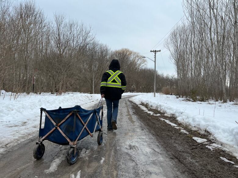 A woman in a black coat with a large high-visibility x on the back pulls a small blue wagon along a muddy path between a snowy, wooded area.