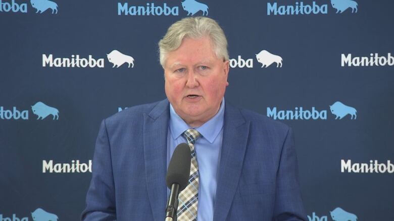 A man in a blue suit jacket and shirt wears a plaid tie in front of a blue Manitoba government background as he speaks into a microphone at a podium.