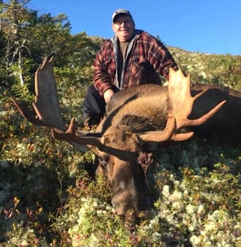 A smiling man in a red and black jacket sits behind a dead moose.