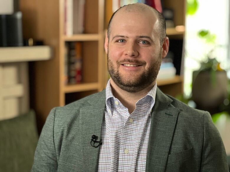 A man standing in an office smiles at the camera. He is wearing a grey blazer and a checkered button-up shirt. There are bookshelves behind him. 