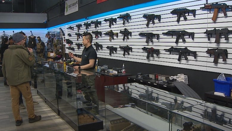 a man talks to a man standing behind a counter. there are rows of airsoft guns lining the wall