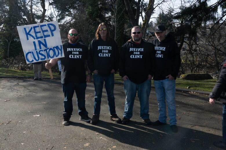 Four men in black hoodies that read 'justice for Clint' stand together in front of trees, with the one at the far left holding a placard that reads 'keep in custody.'