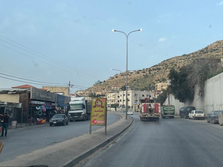 Arabic writing is shown on a road sign with vehicles going in both directions and sandy rocky hilly terrain in the background.