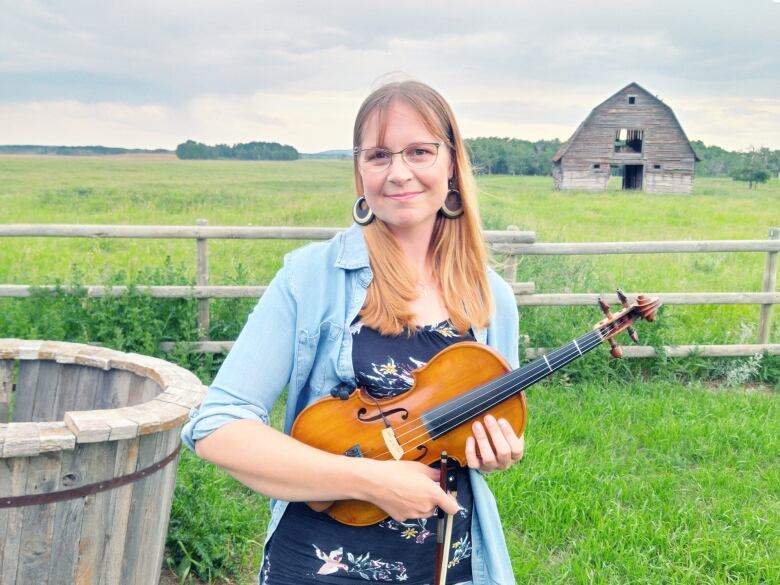 A woman holds a fiddle and stands in a rural scene with a barn behind her.