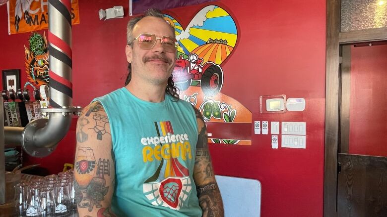 A man in a mullet and glasses leans against a counter. The man is wearing a turquoise blue armless shirt with the phrase 