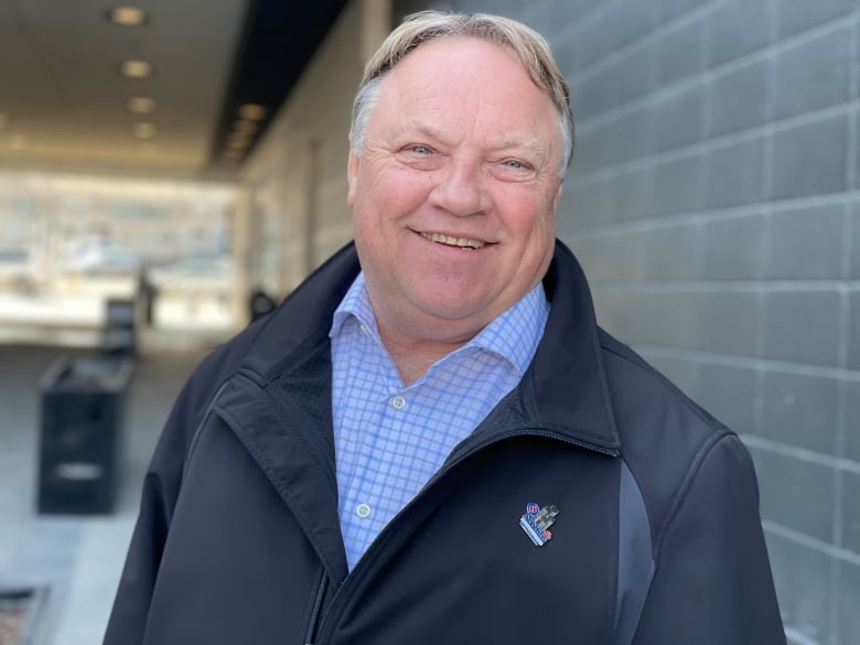 A man wearing a black jacket and blue checked shirt smiles