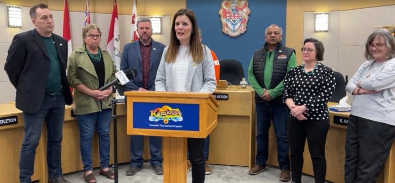 Seven people surround one woman speaking at a podium.