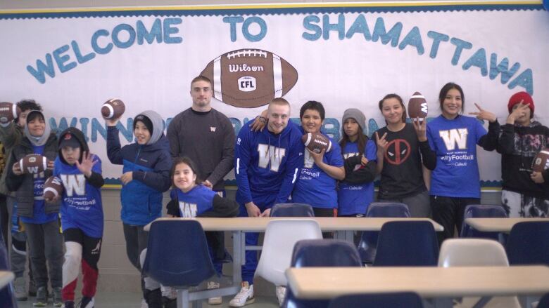 Two football players and several youth stand in front of a chalk board with a giant sign that reads 