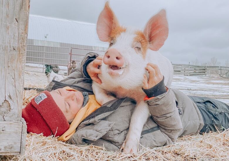 Sanctuary owner, wearing a red beanie, lays on the floor hugging a pig