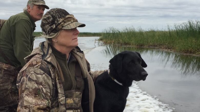 A man and woman in camouflage clothes ride through a wetlands area in a boat along with a black dog.