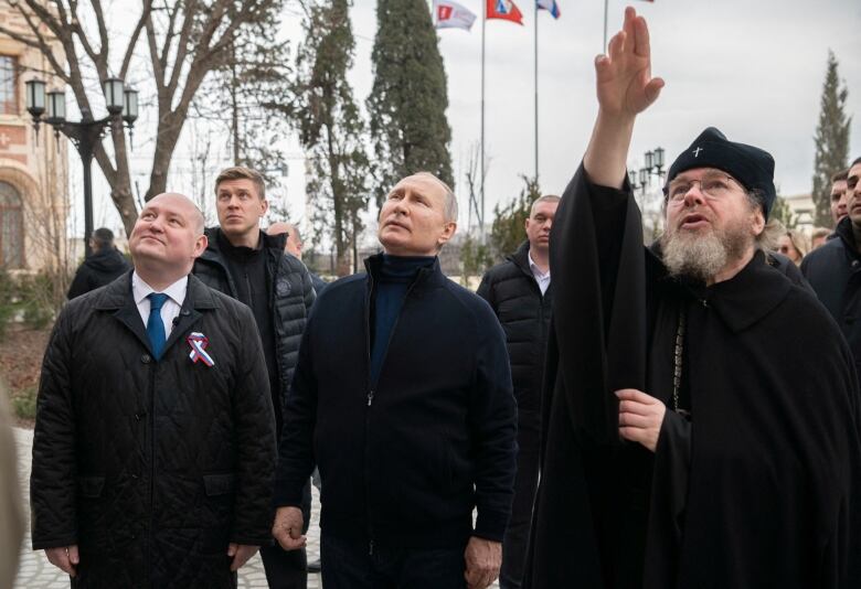 Several men, dressed in black clothing, stand in a group looking upward.  A man on the right, wearing black religious clothing, gestures upward with his hand. 