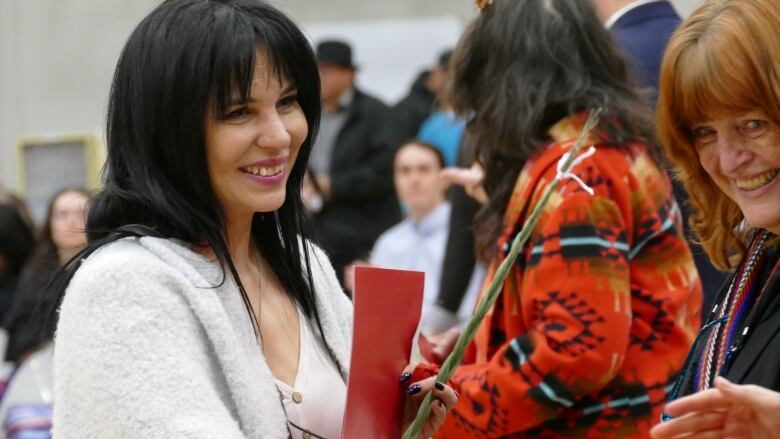 A woman is holding a red folder and a flower and smiling. A hand is reaching in from out of frame, while another woman smiles from the side.