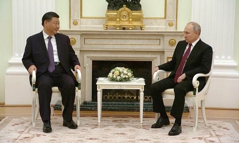 Two men in suits are shown seated in chairs in an ornately decorated room.