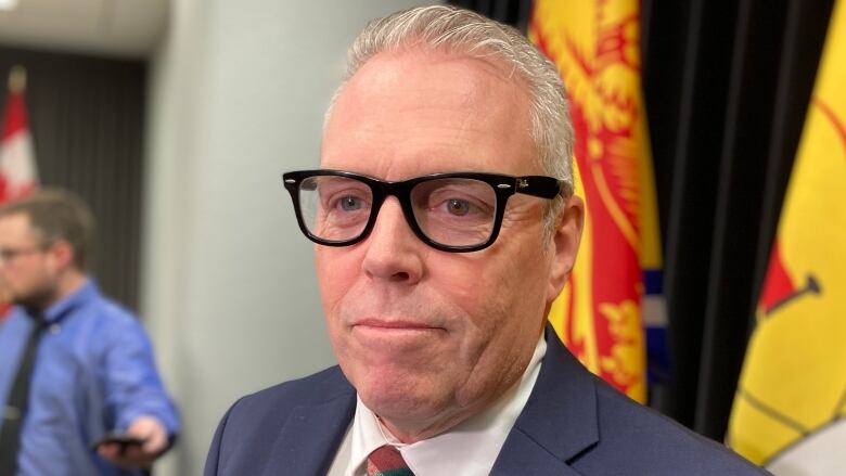 A man with grey hair and glasses wears a blue suit and a tartan tie. 