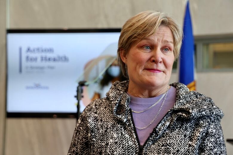 A woman with short blonde hair wearing a black and white jacket with a purple sweater and silver necklace stands in front of a large flat screen TV and a Nova Scotia flag.