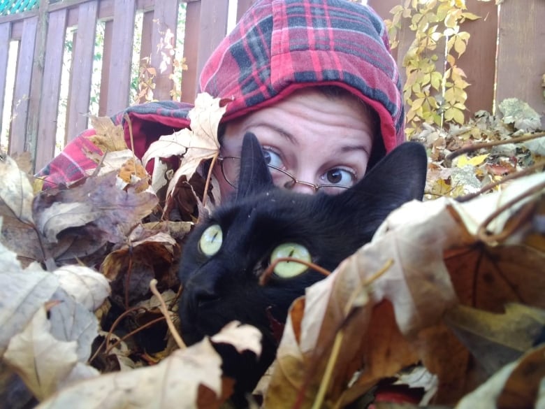 A woman and a cat hide in a pile of leaves.