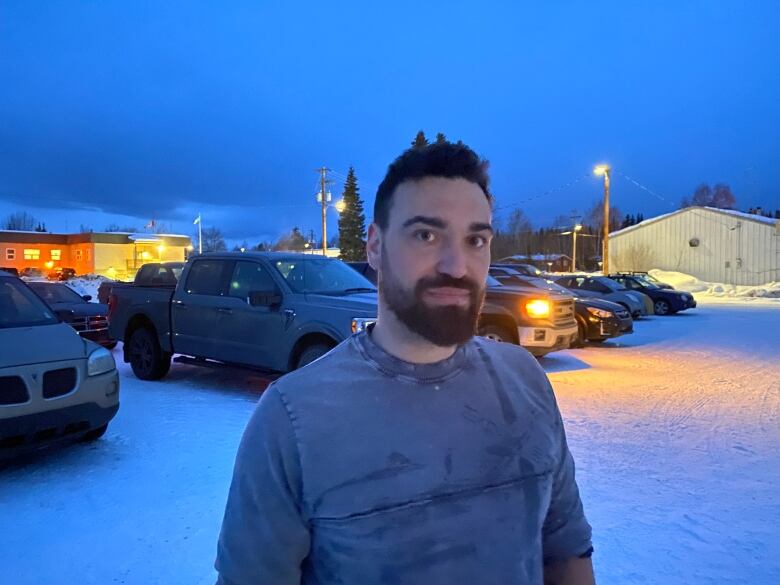 A man with a beard stands in a dimly lit, snowy parking lot.