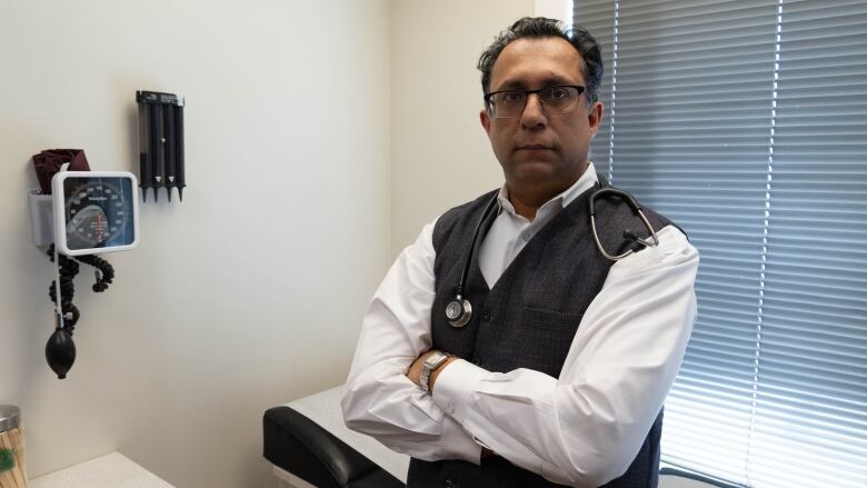 A doctor leans on a bed with his arms crossed.