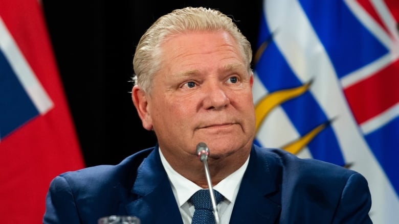 Ontario Premier Doug Ford, wearing a suit and tie, sits in front of small microphone at press conference in front of flags,