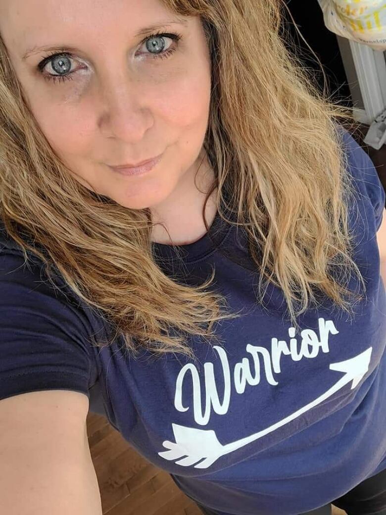 A close-up of a woman with blue eyes and blondish hair, wearing a blue t-shirt that says 