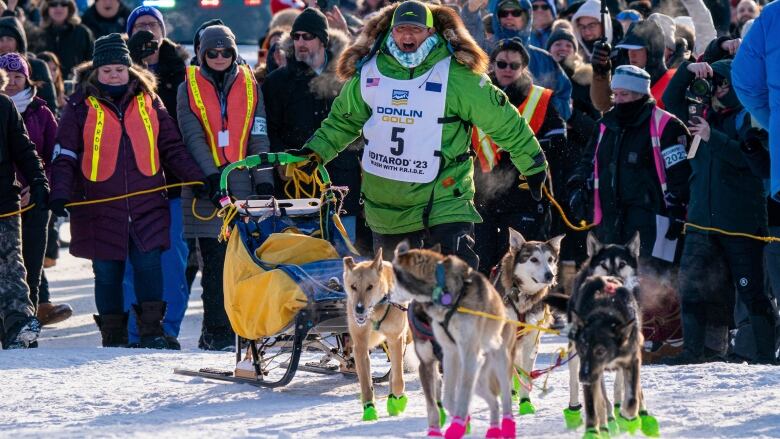 Man in a big parka next to a sled with dogs, in front of a crowd