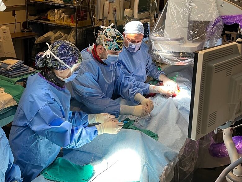 Doctors in blue scrubs hover around a patient on an operating table. The patient is covered in a blue drape.