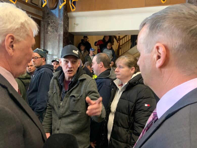 A man with an angry expression on his face points at two men wearing suits. The room they stand in is filled with people.