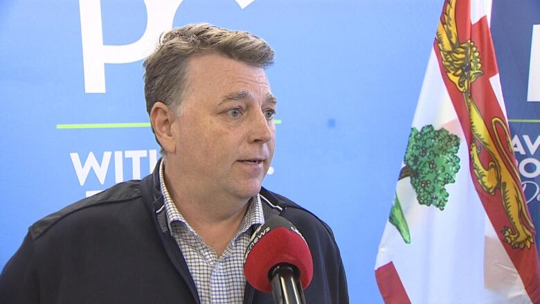 A man talks in front of a microphone. There is a P.E.I. flag and the Progressive Conservative Party logo behind him.