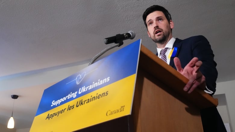 A man in a suit stands at a podium that bears the Ukrainian flag.