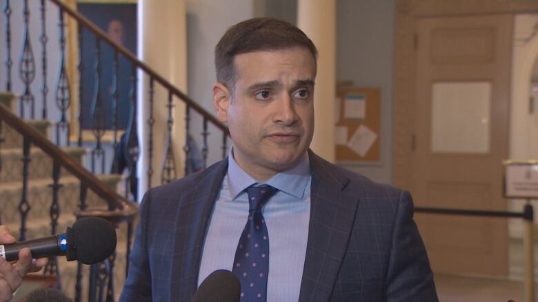 A man wearing a grey blazer with a blue shirt and blue tie stands in front of a staircase.