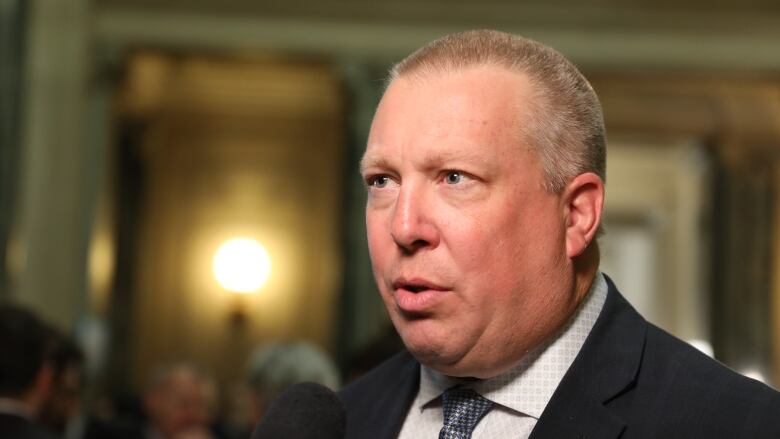 A white man with a buzz cut is wearing a suit with a blue-and-silver tie. He is speaking into a microphone, while standing in a rotunda.