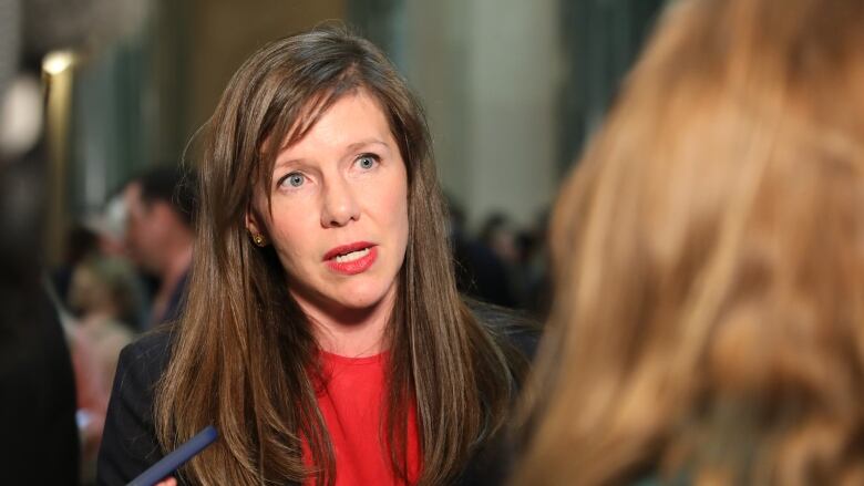 A woman with long brown hair, wearing a black blazer over a red crewneck shirt, speaks to reporters in a rotunda.