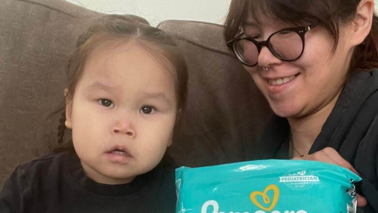 A woman sits with a toddler holding a container of infant formula and a bag of diapers.