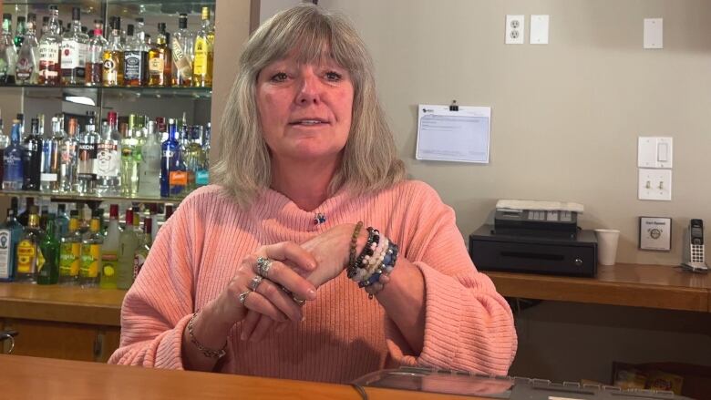 A woman speaks to the camera with alcohol bottles positioned behind her.