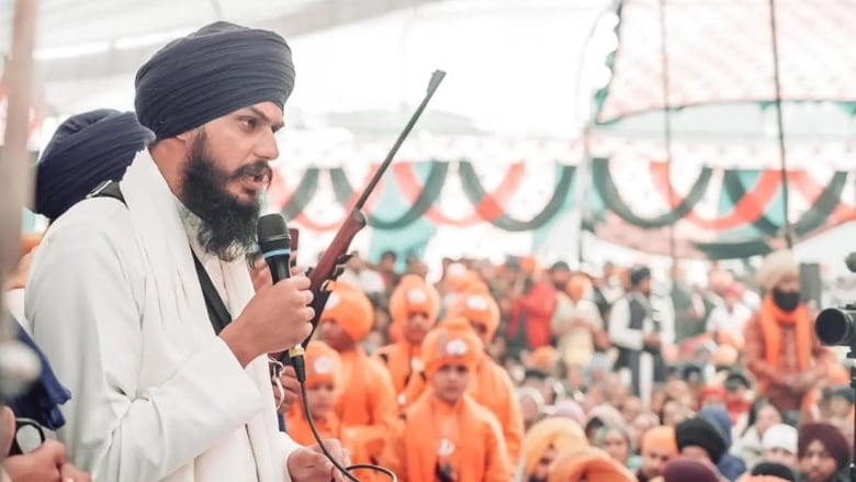 A Sikh man speaks into a mic to a large audience of children and adults under a tented space. A man holds a rifle behind him. 