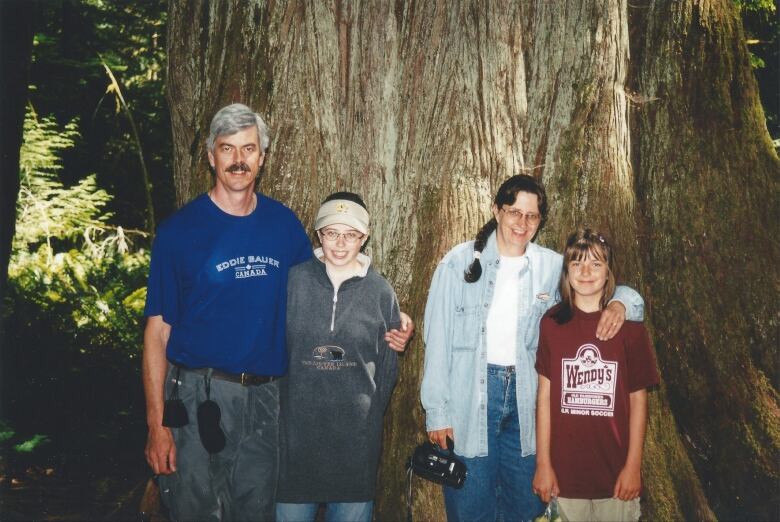 A man with a moustache and grey hair stands in front of a giant tree trunk with two girls and a woman wearing jeans and a jean shirt.