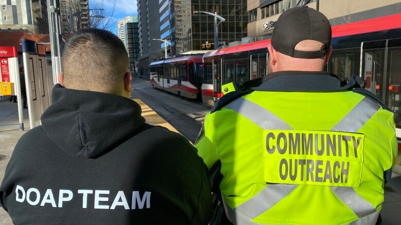 Two men with the words DOAP TEAM and COMMUNITY OUTREACH printed on their backs face away from the camera looking at CTrain tracks in front of them.